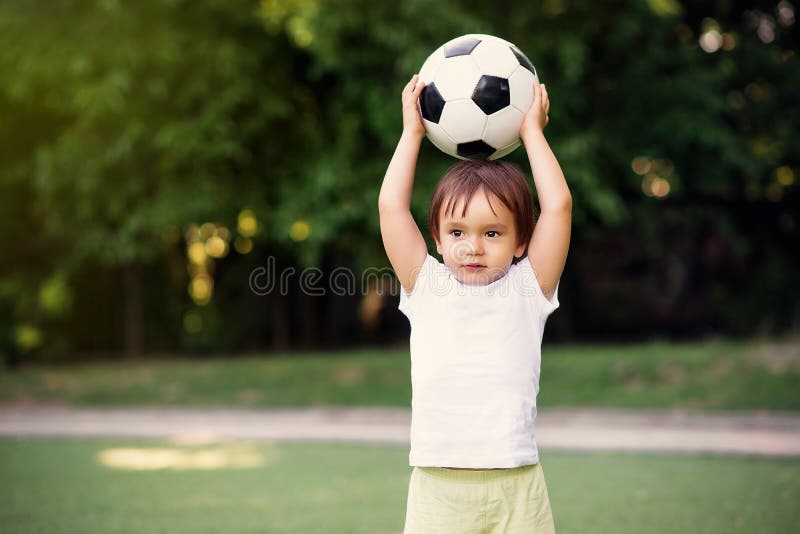 Weinig voetballer bij voetbalgebied in openlucht: de holdingsbal van de peuterjongen boven hoofd klaar om het in zonnige dag te w stock fotografie Weinig voetballer bij voetbalgebied in openlucht: de holdingsbal van de peuterjongen boven hoofd klaar om het in zonnige dag te w stock fotografie