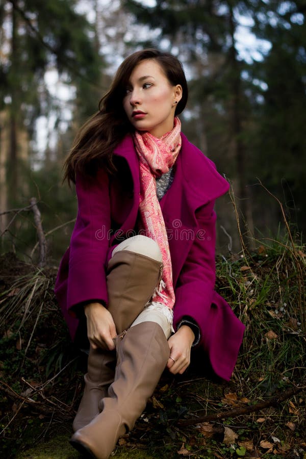 Well-dressed young woman in pink topcoat sitting in forest