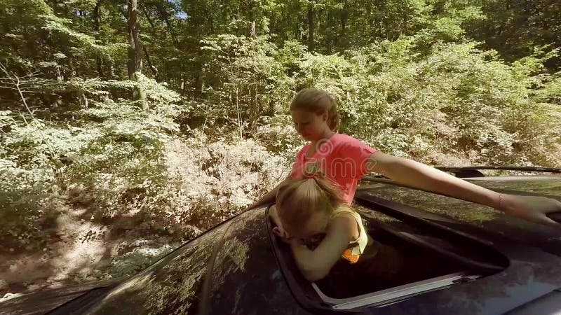 Wellenartig bewegen mit zwei teilt das glückliche lächelnde junge Blondinen ihr von der Luke eines Autos aus, das gegen den Wald