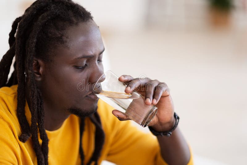 Wellness Concept. Closeup Shot Of Young Black Male Drinking Water From Glass While Relaxing Indoors, African American Man With Dreadlocks Hairstyle Having Refreshing Drink While Relaxing At Home. Wellness Concept. Closeup Shot Of Young Black Male Drinking Water From Glass While Relaxing Indoors, African American Man With Dreadlocks Hairstyle Having Refreshing Drink While Relaxing At Home