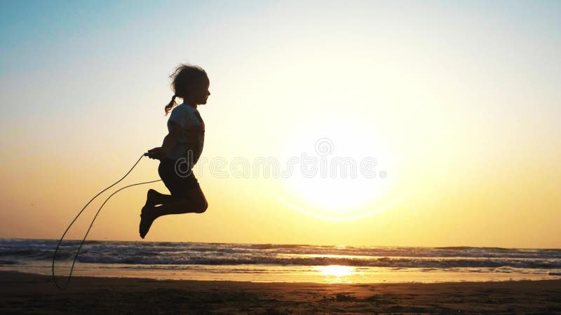 Wenig Mädchen springt auf das Seil auf dem Meersandstrand bei Sonnenuntergang