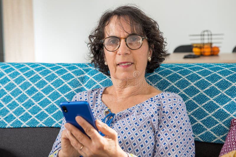 Wesoła brunetka starsza kobieta przy użyciu smartfona siedząc na sofie