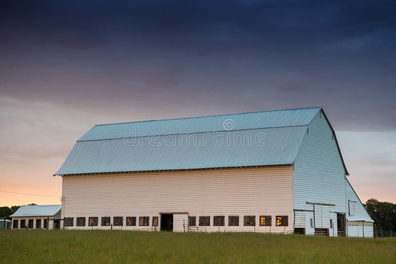 White barn at sunset in a green, grassy field under a sky with purple, yellow, and orange clouds