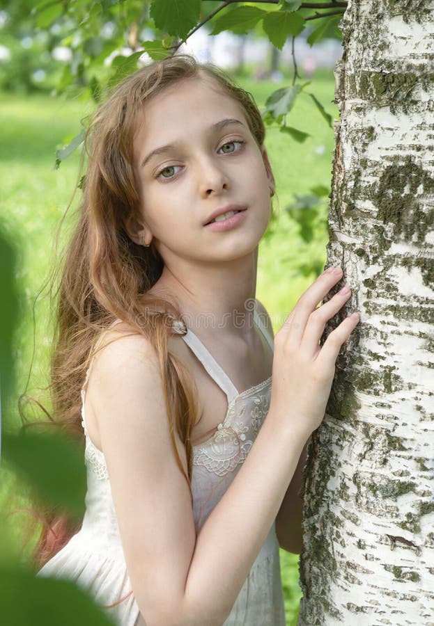 White beautiful girl 11 years old with long hair in a white dress near a birch tree on a green background