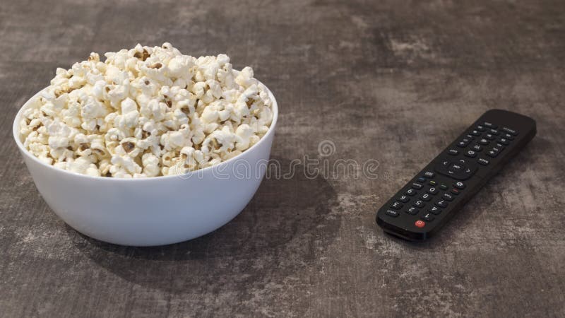 Bowl of popcorn and remote control on a wooden table. White bowl of white salty popcorn and remote control on a wooden table. Angular, close up view stock image Bowl of popcorn and remote control on a wooden table. White bowl of white salty popcorn and remote control on a wooden table. Angular, close up view stock image