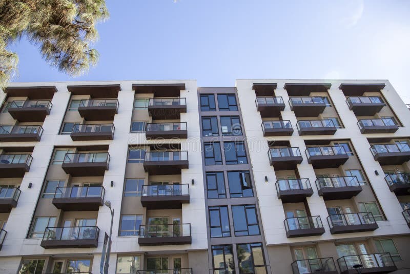 A white and brown apartment building with a glass balconies, lush green trees and a clear blue sky in Los Angeles California