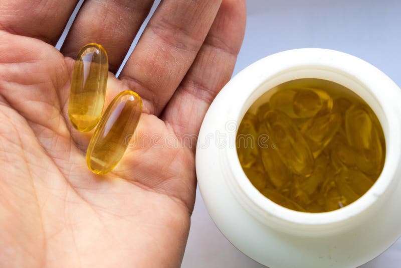 A white can of fish oil in jelly-like transparent yellow capsules and one dose two capsules of capsules in a woman`s palm close-up on a white background. A white can of fish oil in jelly-like transparent yellow capsules and one dose two capsules of capsules in a woman`s palm close-up on a white background