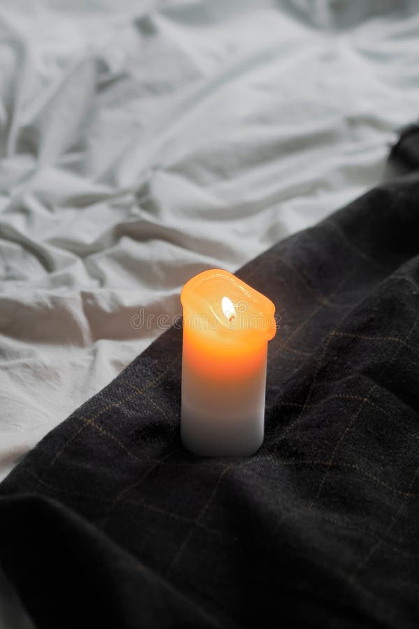 A White Candle with a Lit on a Black Linen Covered Bed Stock Photo