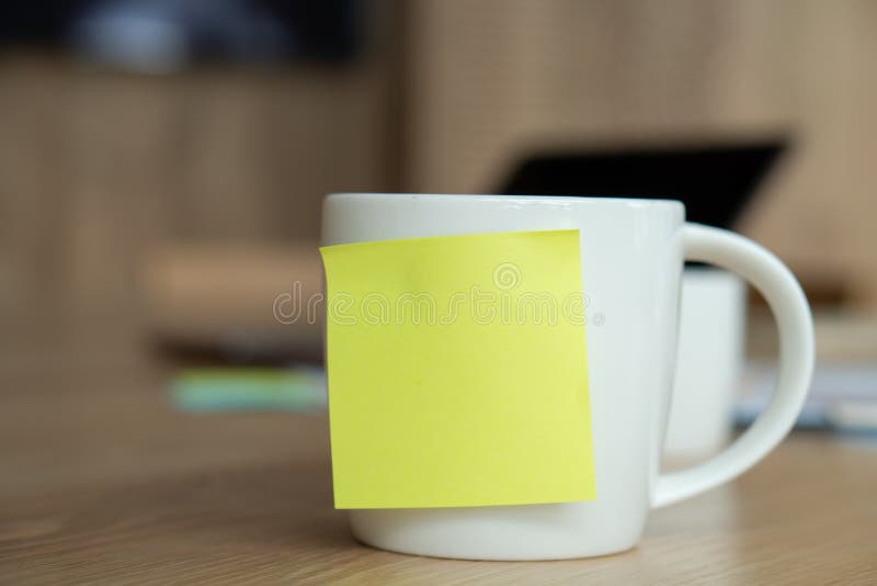 A white coffee cup with a blank yellow post-it on the cup. Resting on the desk and laptop is placed on the back stock image A white coffee cup with a blank yellow post-it on the cup. Resting on the desk and laptop is placed on the back stock image