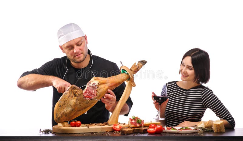 The cook cuts a whole jamon on a wooden stand with a fillet knife , the girl with a glass of wine looks at it. Isolated