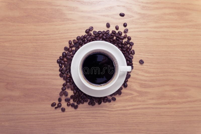 White cup with black, hot coffee standing on brown, roasted beans on wood table