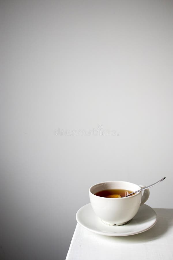 White cup of tea with lemon with a saucer and spoon on a white table. Vertical frame. Empty space