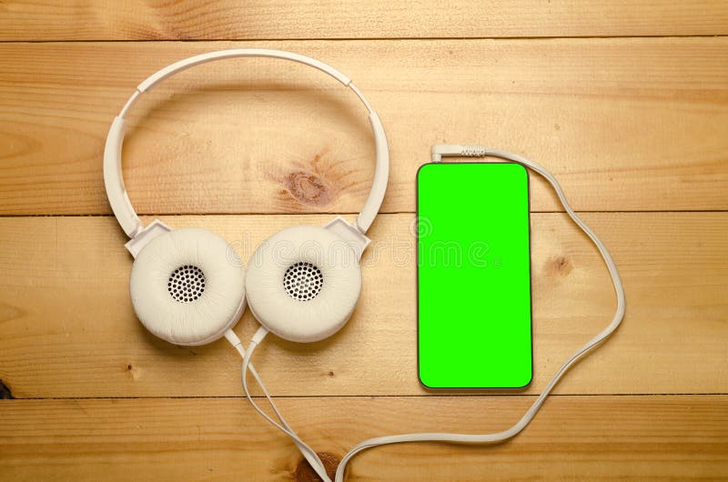 White headphones connected to phone on wooden table. Modern headphone with cable and gadget with green screen