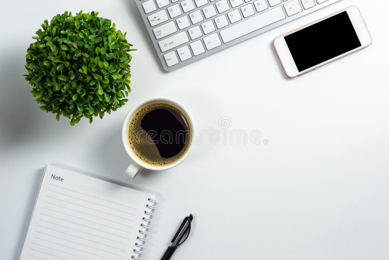 White office desk with black blank screen smartphone, coffee cup, blank notebook, pen, computer keyboard and plant pot