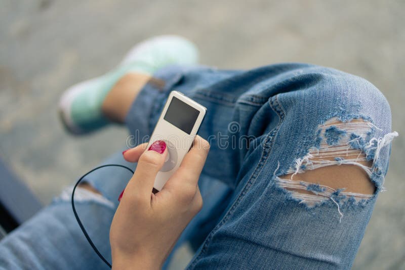 White portable music player in hand with red nail polish. A woman sitting on a bench in the summer. She is dressed in torn jeans and sports shoes.