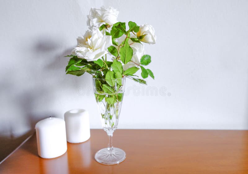 White roses in vase, white candles on wooden commode across white wall. Interior design. Home decor. Copy space.