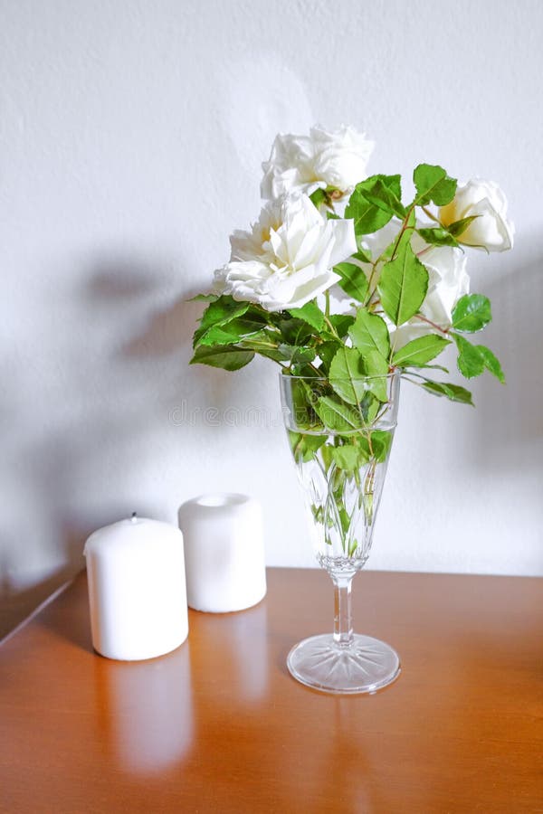 White roses in vase, white candles on wooden commode across white wall. Interior design. Home decor.
