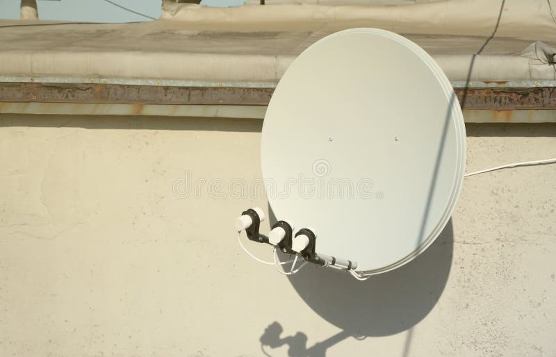 White satellite dish with three converters mounted on residental building rooftop concrete wall. Satellite television advertisement