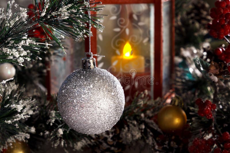 White shiny ball hanging on a branch of a Christmas tree against a red lantern with a candle