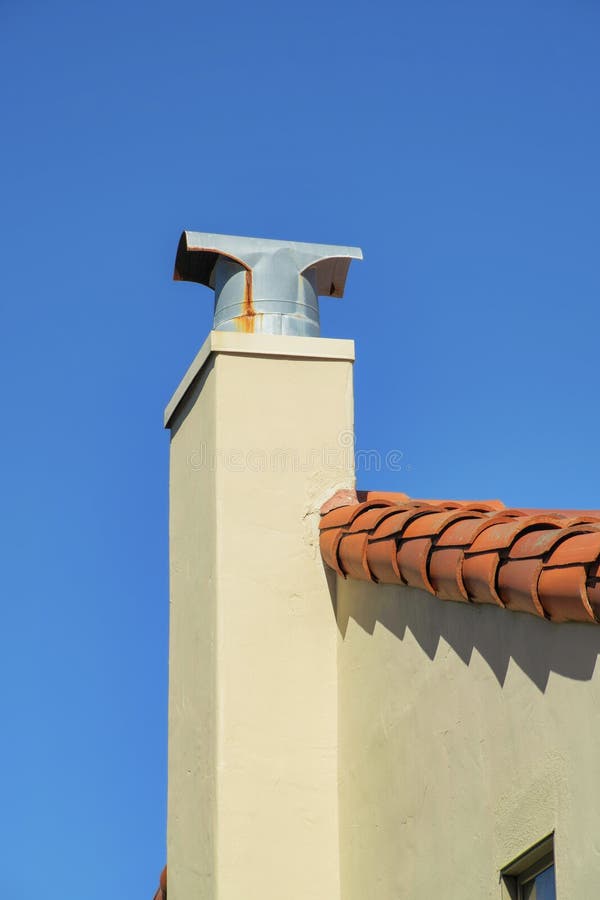 White stucco chimney with metal top with rust marks and red adobe roof tiles on shady roof and house facade in the