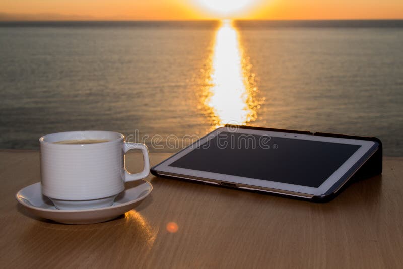 White tablet ipad on table desk with coffee cup during sunset, with sun reflecting on the water