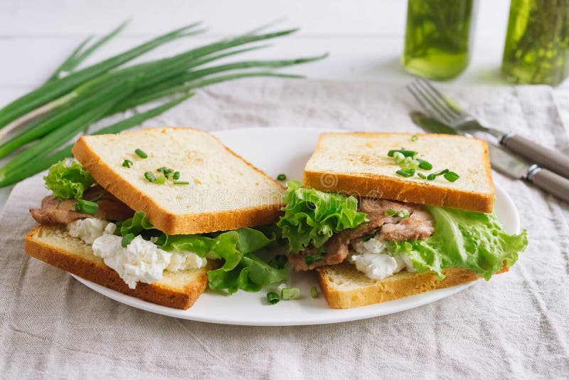 White wheat bread toasted sandwich with pork meat and cheese, salad and sour cream. Chopped chives, bottled olive oil, a knife and