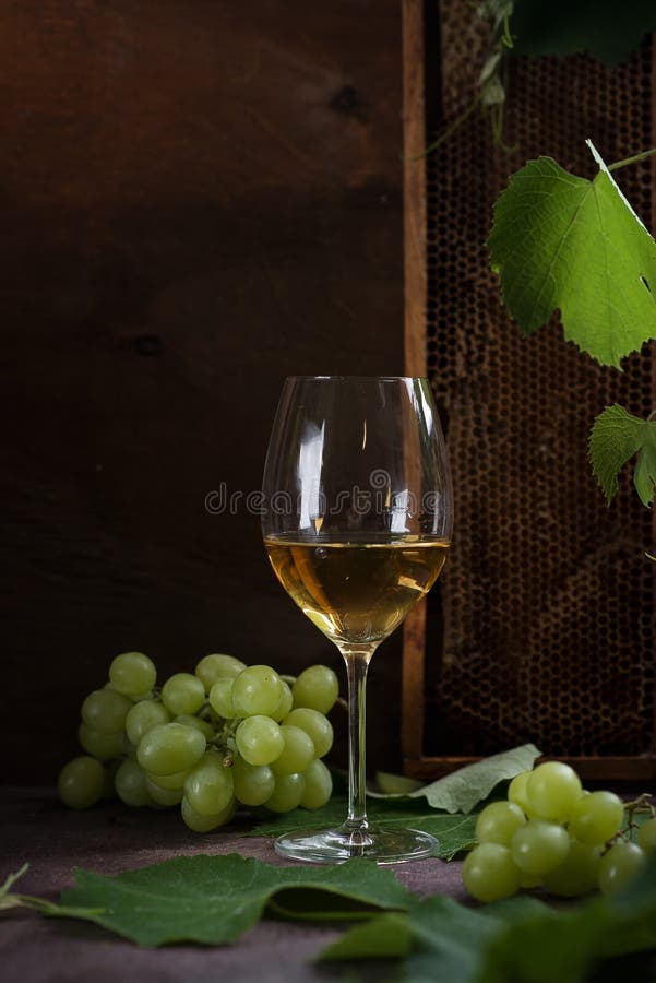 White wine in glasses. Glasses stand on a dark table next to the grape leaves and green grapes. Honeycombs stand in the background