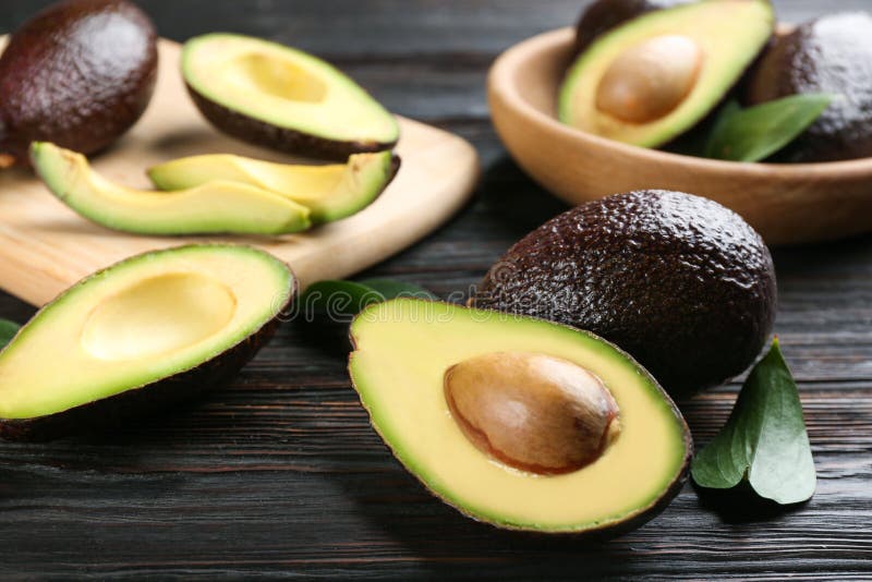 Whole and cut avocados with green leaves on dark wooden table, closeup