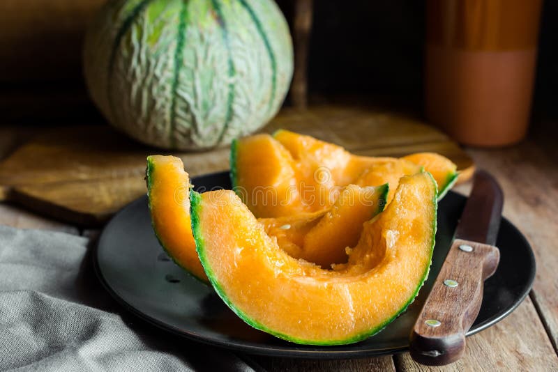 Whole and sliced ripe organic cantaloupe with juicy orange pulp, dark plate, knife, wood cutting board, rustic kitchen interior