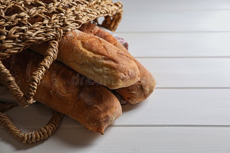 Wicker bag with fresh crispy ciabattas on white wooden table, closeup. Space for text