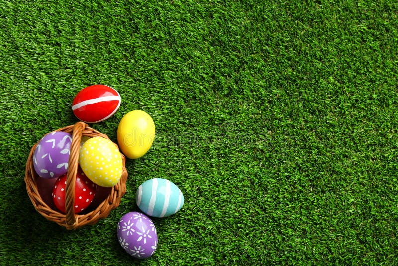 Wicker Basket with Painted Easter Eggs on Green Grass, Top View. Stock