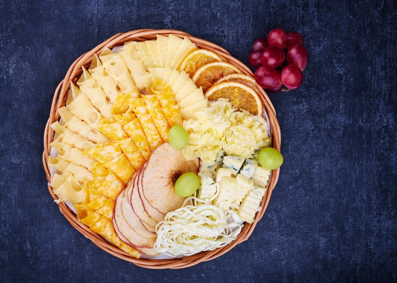 Wicker flat basket with a delicious variety of cheese, dried orange and apple slices on a dark background