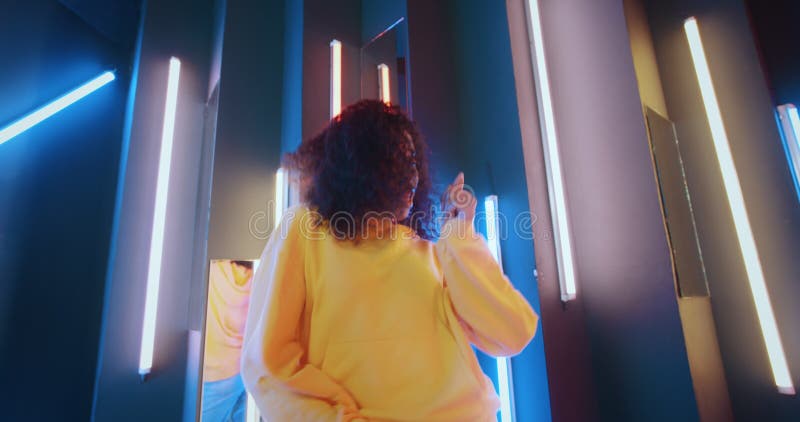 Wide angle positive african american black woman in yellow hoodie dancing in night club backlit with purple and blue. Neon stripes lights. Happy young urban