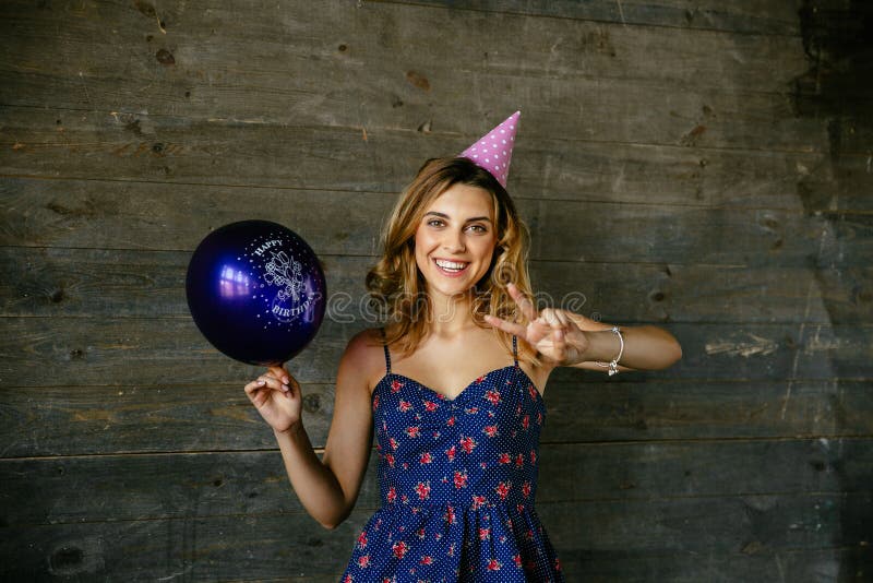 Widely smiling woman in birthday hat celebrating holiday. Cheerful excited girl having fun while celebrating birthday, showing peace sign, holds a balloon royalty free stock images Widely smiling woman in birthday hat celebrating holiday. Cheerful excited girl having fun while celebrating birthday, showing peace sign, holds a balloon royalty free stock images