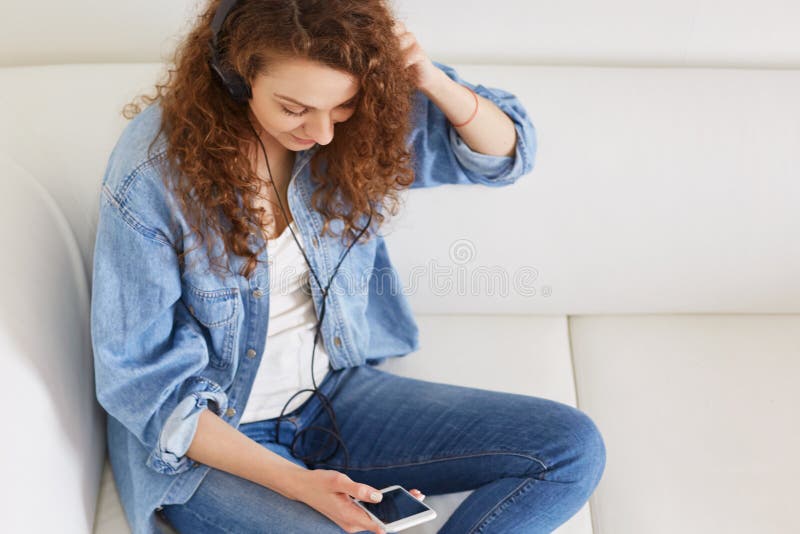 Widok od above uroczy kobieta model ubierający w drelichowej koszula i cajgach, słucha audio ślad przez telefonu komórkowego i hełmofony, ściągają piosenki jej playlistów ludzie rozrywki pojęcie i