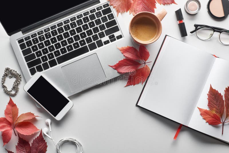 Top view of laptop near smartphone, coffee cup, cosmetics, earphones, glasses, notebook and red leaves of wild grapes on white table,stock image. Top view of laptop near smartphone, coffee cup, cosmetics, earphones, glasses, notebook and red leaves of wild grapes on white table,stock image