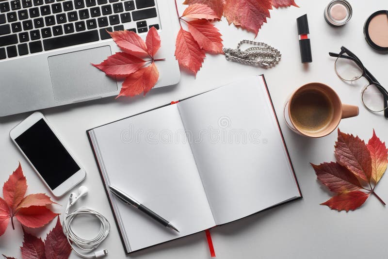 Top view of laptop near smartphone, coffee cup, cosmetics, earphones, glasses, notebook and red leaves of wild grapes on white table,stock image. Top view of laptop near smartphone, coffee cup, cosmetics, earphones, glasses, notebook and red leaves of wild grapes on white table,stock image