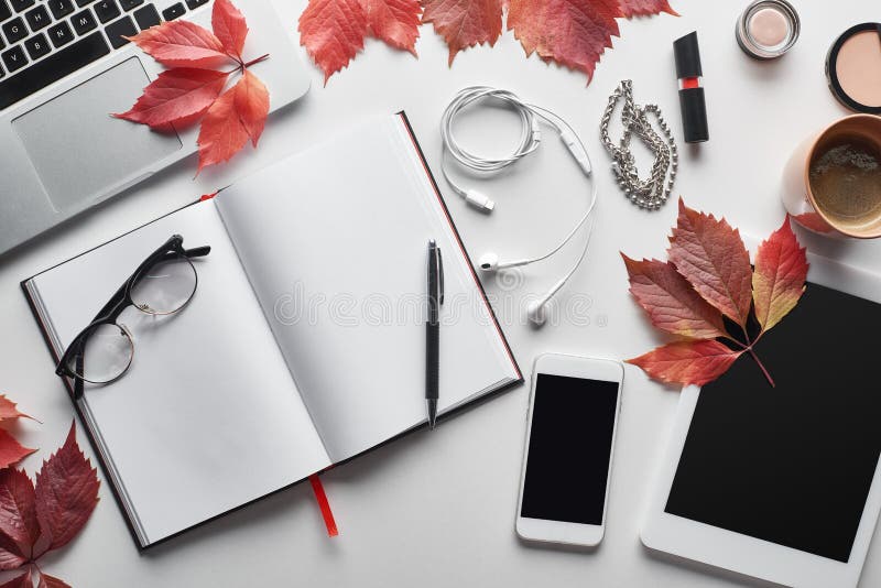 Top view of laptop near smartphone, digital tablet, coffee cup, cosmetics, earphones, glasses, notebook and red leaves of wild grapes on white table,stock image. Top view of laptop near smartphone, digital tablet, coffee cup, cosmetics, earphones, glasses, notebook and red leaves of wild grapes on white table,stock image