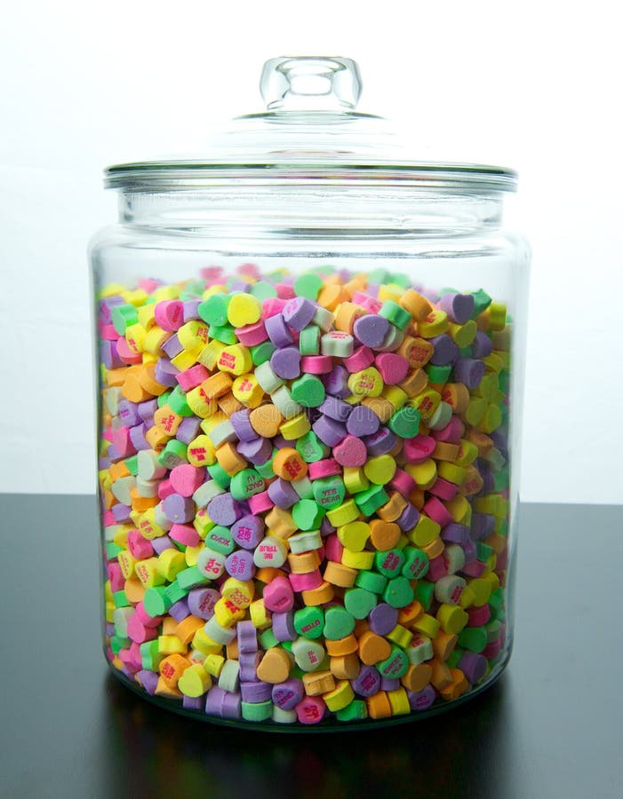 Large glass jar of colorful candy hearts with lid sits on a black table with a white background. Large glass jar of colorful candy hearts with lid sits on a black table with a white background