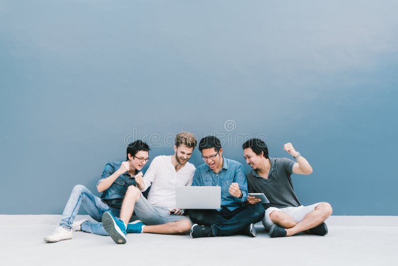 Multiethnic group 4 men celebrate together using laptop computer, sit by blue wall with copy space. College student, information technology gadget, education, social network, or success concept. Multiethnic group 4 men celebrate together using laptop computer, sit by blue wall with copy space. College student, information technology gadget, education, social network, or success concept