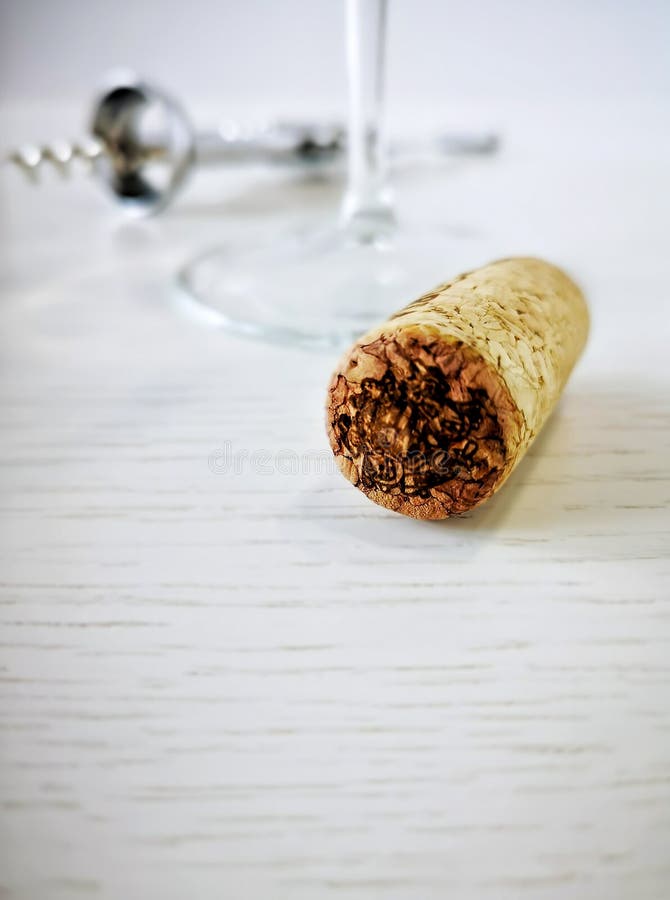 Wine cork is lying on a white wooden table. In the background, a metal corkscrew and an overturned glass with a blue leg