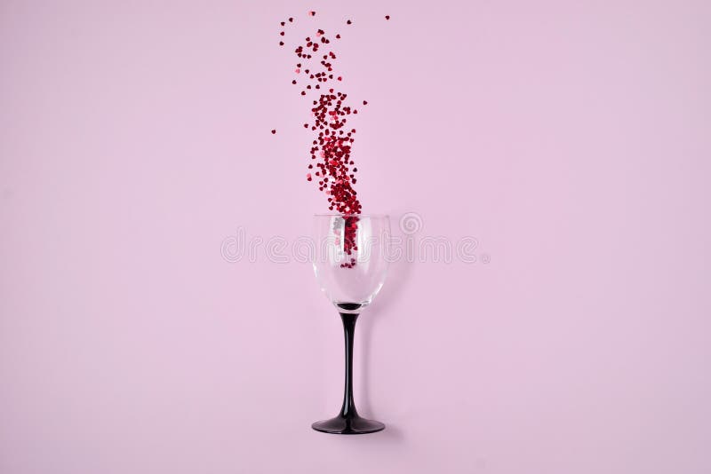 Wine Glass poured out red heart confetti on pink color paper background. Minimal style.