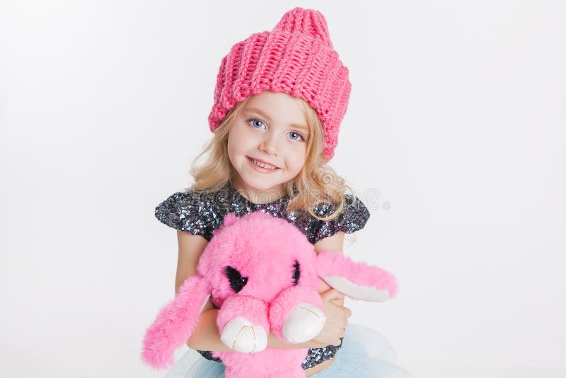 Winter clothes. Portrait of little curly girl in knitted pink winter hat on white. Pink rabbit toy in her hands