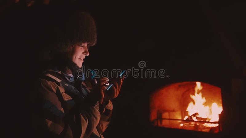 Winter Das Mädchen in einer Jacke und in einem Hut draußen nahe dem Kamin In den Händen, die intelligentes Telefon halten