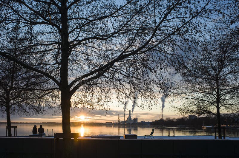 Winter sunset in Vasteras, Sweden. Couple enjoying view of low winter sun reflecting in Malaren lake, bare tree branches against twilight sky, smoke rises vertically in calm weather from plant chimney