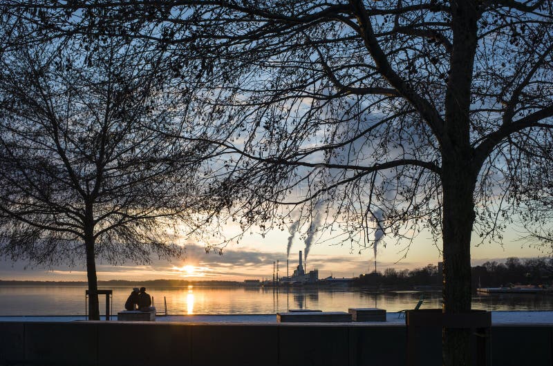 Winter sunset in Vasteras, Sweden. View of low winter sun reflecting in Malaren lake, bare tree branches against twilight sky, kissing couple, smoke rises in calm weather from recycling plant chimney
