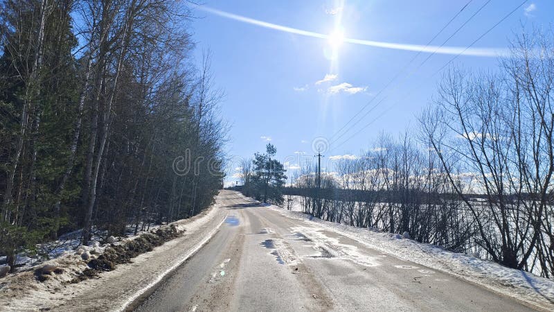 In winter, there is snow on the woods and in the fields. Under the rays of the bright sun, the snow melts on the asphalt road. The royalty free stock photo
