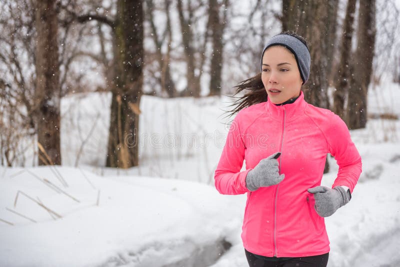 Winter sport fitness girl running in snow wearing wind jacket, gloves, headband and smartwatch. Asian woman healthy and active lifestyle training outside during the cold weather winter season. Winter sport fitness girl running in snow wearing wind jacket, gloves, headband and smartwatch. Asian woman healthy and active lifestyle training outside during the cold weather winter season