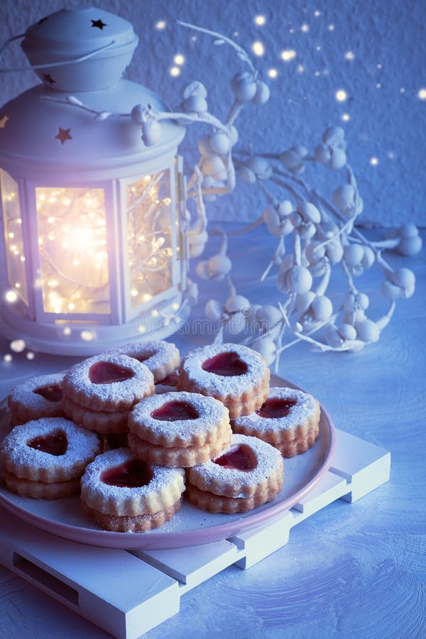Wintertime lantern with lights and Christmas Linzer cookies, biscuits filled with red jam on rustic board.