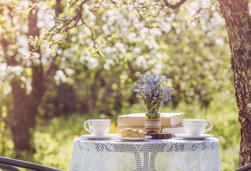 Springtime in garden. Table set with tea cups  boho reuse jar vase with forget me not flowers Myosotis and small stack of books. Summer relax readings concept. Springtime in garden. Table set with tea cups  boho reuse jar vase with forget me not flowers Myosotis and small stack of books. Summer relax readings concept.